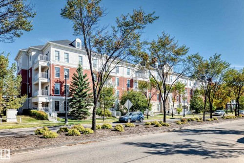 Multi-story building featuring brick and stucco facade, balconies with metal railings, dormer windows, and a landscaped street frontage - #307 1003 Gault Bv Nw, Edmonton, AB 