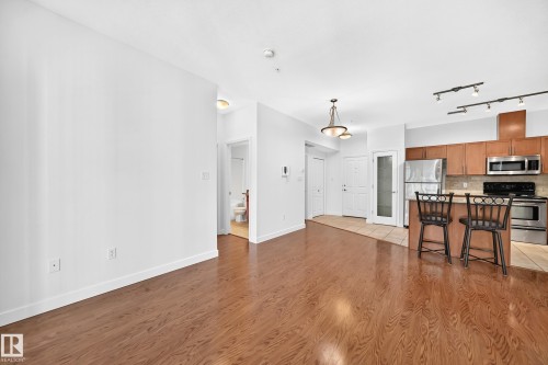 Wood-finish flooring throughout, kitchen with stainless steel appliances, wood cabinetry, and a bar-height counter, tile flooring in the entry and kitchen area, and a semi-flush mount light fixture - #307 1003 Gault Bv Nw, Edmonton, AB 