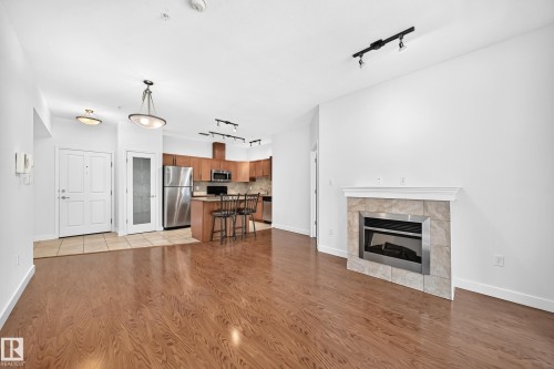 Open-concept living space with wood-finish flooring, a tile-surround fireplace, and a kitchen featuring stainless steel appliances and wood cabinetry - #307 1003 Gault Bv Nw, Edmonton, AB 