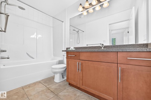 Bathroom featuring a vanity with wood-finish cabinetry, a stone countertop, an integrated sink, a full-width mirror, and a shower-over-tub combination with a white surround - #307 1003 Gault Bv Nw, Edmonton, AB 