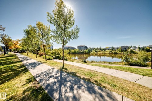 Paved pathways bordering a reflective pond and green spaces, surrounded by mature trees and distant residential structures - #307 1003 Gault Bv Nw, Edmonton, AB 