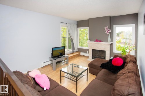 Living area with wood-finish flooring, a stone-clad fireplace, and multiple windows - #407 9810 178 St Nw, Edmonton, AB 