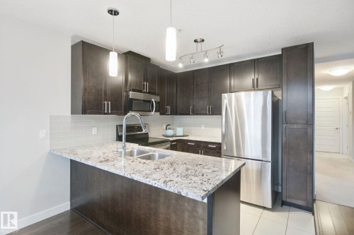 Kitchen featuring dark wood-finish cabinetry, stainless steel appliances, light-toned countertops, and a subway tile backsplash - #125 5151 Windermere Bv Sw Sw, Edmonton, AB 