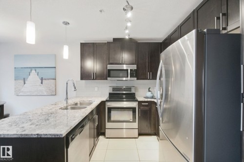 Kitchen featuring dark wood-finish cabinetry, light-toned countertops, stainless steel appliances, and a tile backsplash - #125 5151 Windermere Bv Sw Sw, Edmonton, AB 
