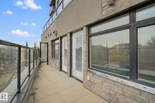 Balcony featuring a glass railing, exterior sconce lighting, stone veneer wall, and two access doors - #125 5151 Windermere Bv Sw Sw, Edmonton, AB 