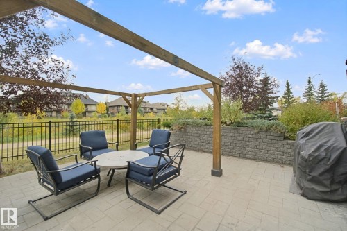 Paved patio area featuring a wooden pergola, black metal fencing, and a tiered retaining wall with integrated landscaping - #125 5151 Windermere Bv Sw Sw, Edmonton, AB 