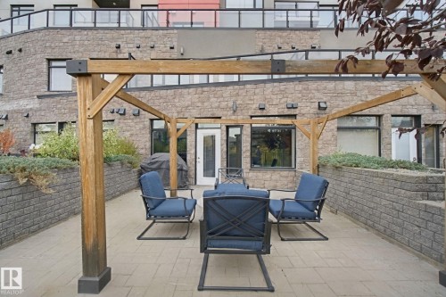 Paved outdoor patio featuring a wooden pergola, raised planting beds with retaining walls, and a stone facade - #125 5151 Windermere Bv Sw Sw, Edmonton, AB 