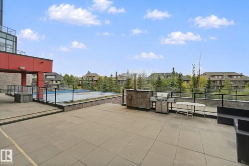 Expansive tiled patio featuring a stone-clad outdoor kitchen, stainless steel grill, and glass panel railings - #125 5151 Windermere Bv Sw Sw, Edmonton, AB 