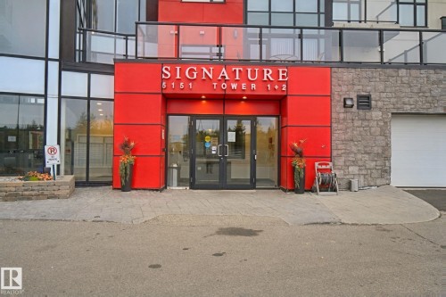 Commercial building entrance featuring prominent red paneling, stone facade accents, a glass double-door entry, and multiple large windows - #125 5151 Windermere Bv Sw Sw, Edmonton, AB 