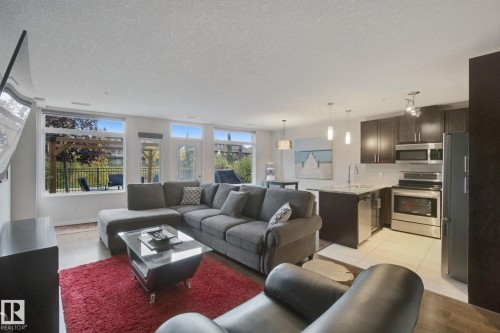 Open concept living space with wood-finish flooring, a kitchen island, stainless steel appliances, and a private patio with a pergola - #125 5151 Windermere Bv Sw Sw, Edmonton, AB 