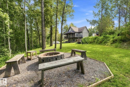 Gravel patio featuring a stone-lined fire pit, surrounded by multiple wooden benches, all set within a landscaped yard with a sloped, treed backdrop - #45 53122 Rge Road 14, Rural Parkland County, AB 