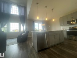 Open-concept living space featuring wood-finish flooring, a kitchen island with integrated sink, stainless steel appliances, and a two-story window array - 