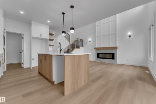 Open-concept living space with wood-finish flooring, a contemporary fireplace with a stone surround and wood mantel, and a kitchen island with pendant lighting - 794 Bank Drive, Leduc, AB 