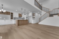 Open-concept living area featuring wood-finish flooring, a contemporary kitchen with white and wood-tone cabinetry, a central island, and a staircase with open risers and metal balusters - 