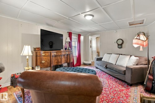 Finished basement recreation room featuring drop ceiling with recessed lighting, wood-finish dresser with brass hardware, flat-screen television mounted above, upholstered seating, and layered area rugs over wood flooring - 12707 73 Street Nw, Edmonton, AB 