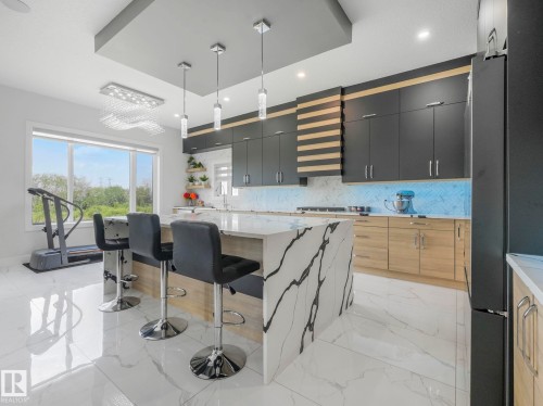 Modern kitchen featuring a large center island with waterfall countertop, recessed ceiling details, pendant lighting, built-in cabinetry, and tile flooring - 120 38 Street Sw, Edmonton, AB 