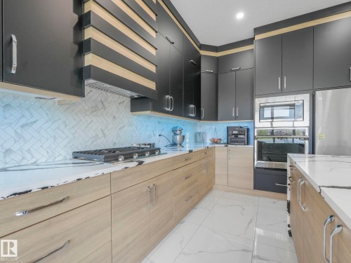 Contemporary kitchen featuring dark flat-panel cabinetry, light wood-finish lower cabinetry, white stone countertops, herringbone-patterned tile backsplash, and stainless steel appliances - 120 38 Street Sw, Edmonton, AB 