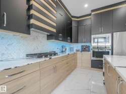 Contemporary kitchen featuring dark flat-panel cabinetry, light wood-finish lower cabinetry, white stone countertops, herringbone-patterned tile backsplash, and stainless steel appliances - 