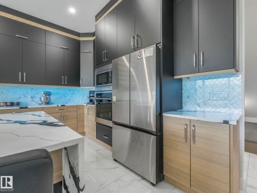 Contemporary kitchen featuring two-tone cabinetry, stainless steel appliances, a large center island with a waterfall countertop, and a herringbone-patterned tile backsplash - 120 38 Street Sw, Edmonton, AB 
