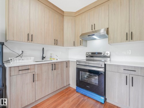 Light wood-finish cabinetry with black hardware, white countertops, stainless steel range with matching hood vent, and a black matte faucet - 120 38 Street Sw, Edmonton, AB 
