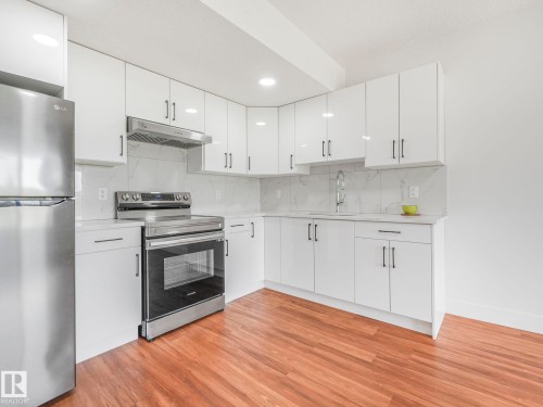 Modern kitchen featuring white cabinetry, stainless steel appliances, wood-finish flooring, white countertops, and a white tile backsplash - 120 38 Street Sw, Edmonton, AB 