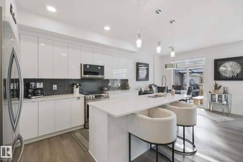 Contemporary kitchen featuring a large island with countertop seating, high-gloss white cabinetry, stainless steel appliances, and a dark tiled backsplash - 153 Cavanagh Common Sw, Edmonton, AB 