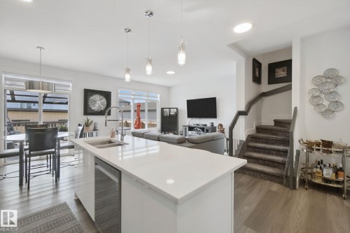 Kitchen island with a white countertop and integrated sink, stainless steel dishwasher, wood-finish flooring, recessed lighting, and a staircase with carpeted treads - 153 Cavanagh Common Sw, Edmonton, AB 