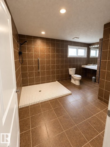 Spacious bathroom featuring full wall and floor tiling, a walk-in shower with a mosaic tile base, a floating vanity with a single sink, and dual windows - 18929 99A Avenue Nw, Edmonton, AB 