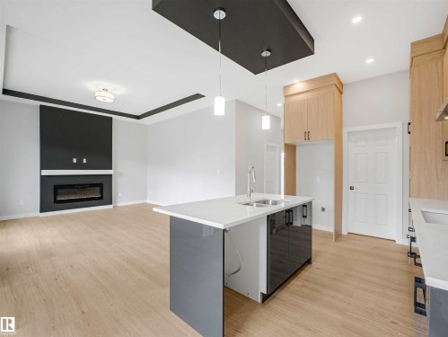 Light wood-finish flooring throughout, kitchen island with integrated sink, light wood cabinetry, recessed ceiling lighting, and a built-in fireplace - 22 Dalquist Bay, Leduc, AB 