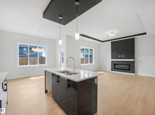 Kitchen island with integrated double basin sink, light-toned wood-finish flooring, recessed ceiling with accent lighting, contemporary linear fireplace with mantel, and multiple windows providing ample natural light - 22 Dalquist Bay, Leduc, AB 