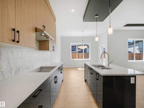 Kitchen featuring wood-finish cabinetry, hexagonal tile backsplash, light-toned countertops, and a center island with a double basin sink and stainless steel faucet - 22 Dalquist Bay, Leduc, AB 