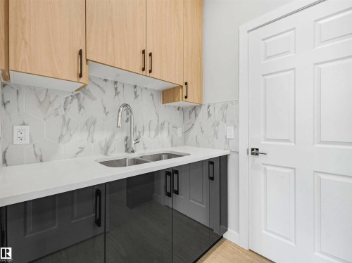 Dual-tone cabinetry with light wood-finish upper cabinets and high-gloss gray lower cabinets, undermount stainless steel sink with a high-arc faucet, and a patterned tile backsplash, all complemented by a solid surface countertop - 22 Dalquist Bay, Leduc, AB 