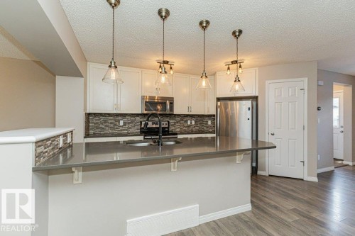 Kitchen featuring white cabinetry, stainless steel appliances, a multi-tone tile backsplash, and a dark countertop island with pendant lighting - 3203 Carpenter Landing Sw, Edmonton, AB 