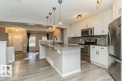Kitchen featuring an eat-at island with pendant lighting, stainless steel appliances, white cabinetry, and a mosaic tile backsplash - 3203 Carpenter Landing Sw, Edmonton, AB 