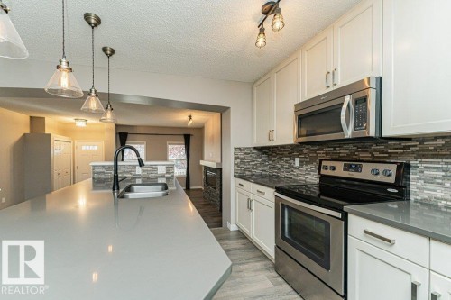 Expansive island with integrated sink and matte black faucet, pendant lighting, stainless steel appliances, white cabinetry with contemporary hardware, and mosaic tile backsplash - 3203 Carpenter Landing Sw, Edmonton, AB 