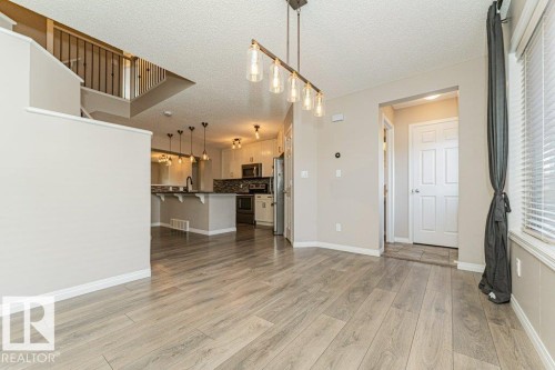 Spacious living area featuring wood-finish flooring, a contemporary linear chandelier, and a bright window with blinds - 3203 Carpenter Landing Sw, Edmonton, AB 