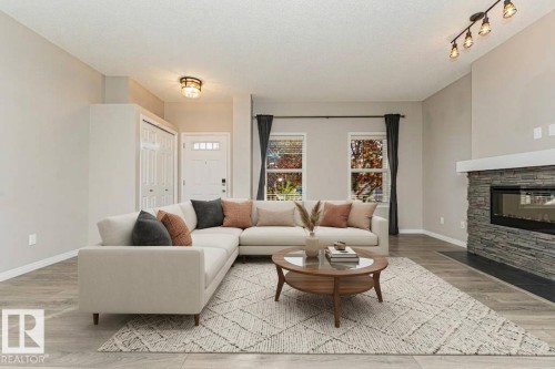 Wood-finish flooring, stone-clad fireplace with a hearth, dual windows with dark drapery, and a white entry door with frosted window panels - 3203 Carpenter Landing Sw, Edmonton, AB 