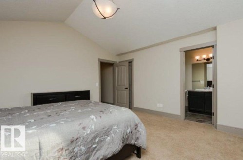 Vaulted ceiling, carpet flooring, built-in black headboard, black bathroom vanity with single sink, and three-light vanity fixture - 7233 Armour Crescent Sw, Edmonton, AB 
