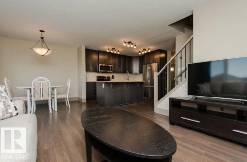Open-concept living space featuring wood-finish flooring, a kitchen with dark cabinetry, stainless steel appliances, and a staircase with wrought iron balusters - 7233 Armour Crescent Sw, Edmonton, AB 