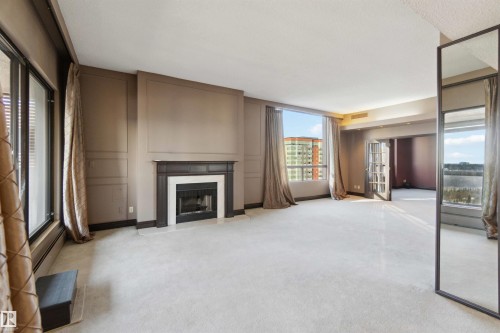 Spacious room featuring carpeting, a fireplace with a dark wood mantel, extensive window treatments, and French doors - #12A 10050 118 St Nw, Edmonton, AB 
