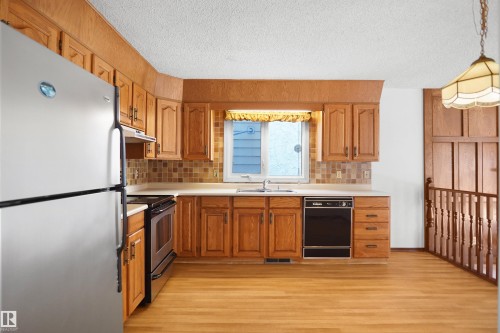 Kitchen featuring wood cabinetry, light-toned countertops, a stainless steel refrigerator, a black dishwasher, and wood-finish flooring - 12311 25 Avenue Nw, Edmonton, AB 