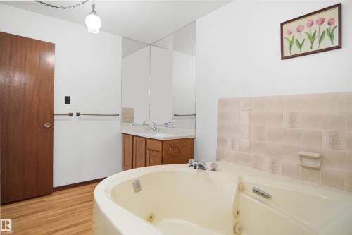 Jetted bathtub, wood-finish flooring, wood vanity with dual sinks, wall-mounted mirror, and tiled bath surround - 12311 25 Avenue Nw, Edmonton, AB 
