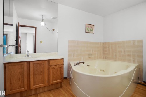 Corner jetted tub with tiled surround, wood-finish flooring, and a vanity with a single sink and wood cabinetry - 12311 25 Avenue Nw, Edmonton, AB 