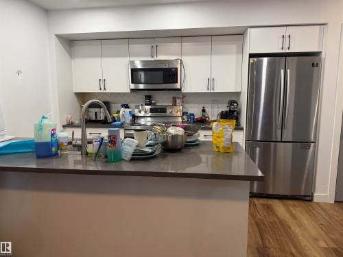 Stainless steel appliances, white cabinetry with black hardware, tile backsplash, wood-finish flooring, and a kitchen island with a built-in sink - 11442 82 Street Nw, Edmonton, AB 