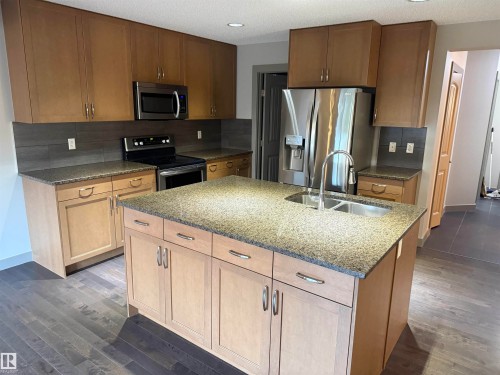 Kitchen featuring a central island with a double basin sink, stainless steel appliances, wood-finish cabinetry, granite countertops, and dark wood-finish flooring - 7065 Cardinal Way Sw, Edmonton, AB 