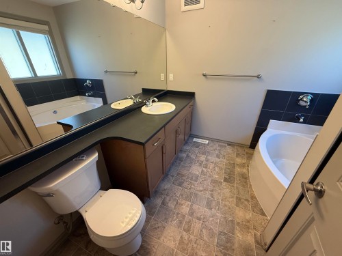 Dual vanity with dark countertops, wood-finish cabinetry, and a large wall-mounted mirror - 7065 Cardinal Way Sw, Edmonton, AB 