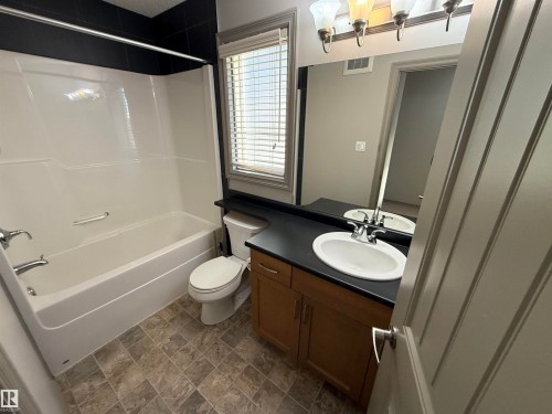White bathtub with chrome fixtures, black tiled shower surround, wood-finish vanity with a single basin sink, and a large wall-mounted mirror - 7065 Cardinal Way Sw, Edmonton, AB 