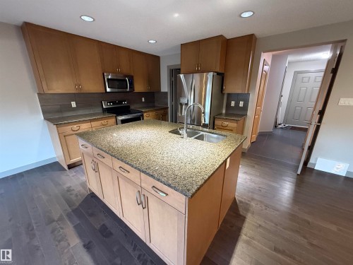 Kitchen featuring an island with a dual-basin sink, wood-finish cabinetry, stainless steel appliances, granite countertops, and wood-finish flooring - 7065 Cardinal Way Sw, Edmonton, AB 