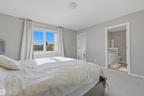 Bright bedroom featuring a large window, light gray carpeting, and direct access to a bathroom with a vanity and toilet - 3151 Trelle Loop Nw, Edmonton, AB 