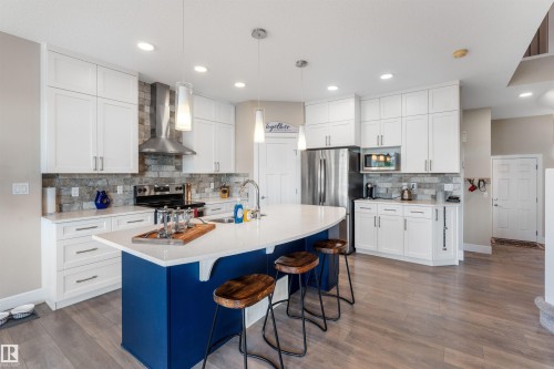 Kitchen featuring a large central island with a curved countertop, white cabinetry, integrated stainless steel appliances, wood-finish flooring, and a stone tile backsplash - 3334 Parker Loop Sw, Edmonton, AB 